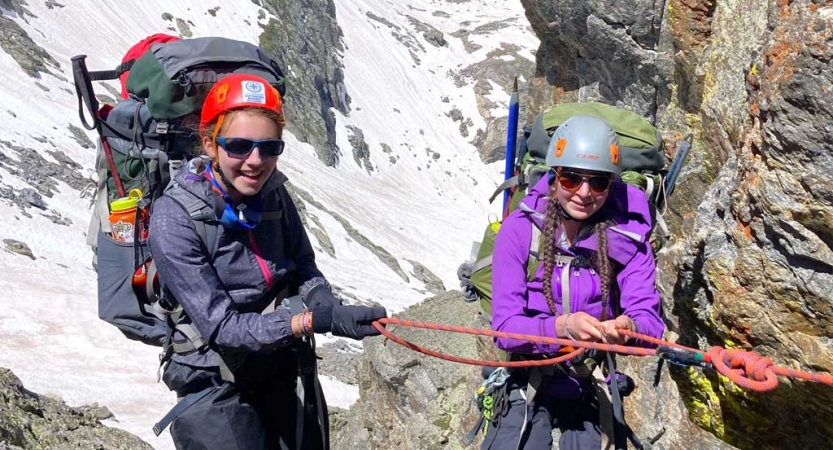 two people wearing safety gear and backpacks hold onto a rope in front of a mountainous snowy landscape